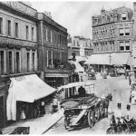 Shops with awnings and two storeys above them; horse drawn carts in street.