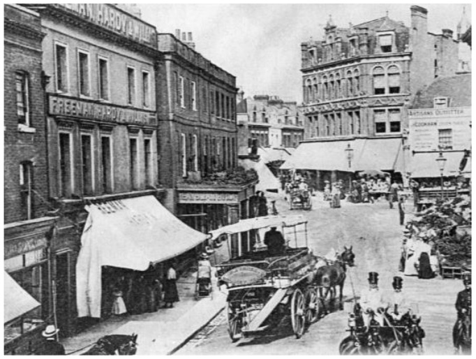 Shops with awnings and two storeys above them; horse drawn carts in street.